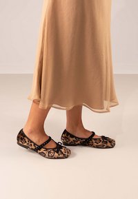 Bailarinas de tela de estampado de leopardo, con una correa negra con adornos y un lazo en la punta, suela plana. Llevadas con un vestido fluido beige.