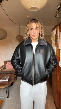 Giacca bomber in pelle nera con colletto grigio peloso, dotata di cerniera frontale e logo in rilievo sul petto. Indossata sopra una maglietta bianca.