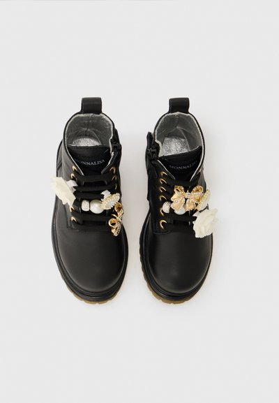 Botas de cuero negras hasta el tobillo con ojales dorados y flores blancas decorativas, perlas y lazos adheridos a los cordones, vistas desde arriba.