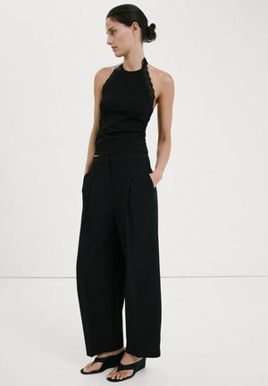 Ženska s temnimi lasmi, spetimi v figo, oblečena v črno brezrokavnik z vrvico okoli vratu, široke ohlapne črne hlače z razširjenimi hlačnicami in črne klini sandale, stoji z rokami v žepih.