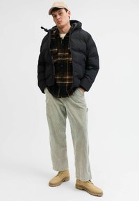 Svart dunjakke med hette over en mørk rutete skjorte. Lysegrønne kordfløyelbukser og beige snørebukser fullfører antrekket.