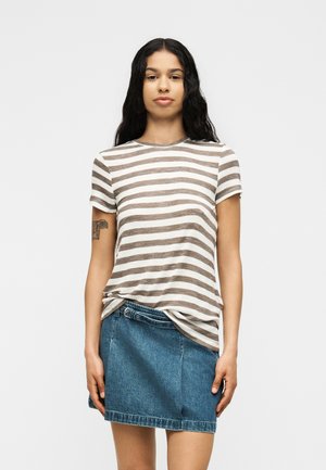 Femme aux longs cheveux foncés portant un t-shirt à manches courtes rayé marron et blanc, rentré dans une jupe en denim bleu, sur un fond uni.