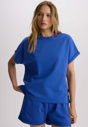 Niebieski bawełniany t-shirt z podwiniętymi rękawami i luźnym krojem, w połączeniu z dopasowanymi niebieskimi szortami. Prosty design, gładka faktura, bez wyraźnych wzorów.