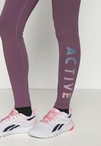 Fioletowe legginsy z napisem "ACTIVE" w niebieskim i różowym kolorze. Materiał jest elastyczny z widocznymi szwami. Noszone z białymi trampkami z różowymi akcentami.
