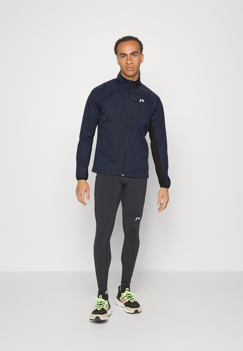 Marineblaue leichte Jacke mit Reißverschluss, schwarzen Akzenten, langen Ärmeln, kombiniert mit schwarzen Leggings und Turnschuhen mit neongrünen Schnürsenkeln.