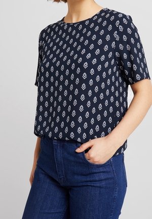 Femme portant un chemisier bleu marine à manches courtes avec des motifs géométriques blancs en forme de losange, rentré dans un jean taille haute bleu foncé.