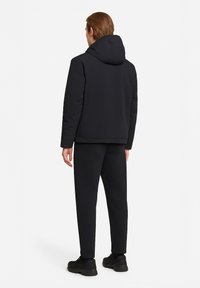 Giacca nera imbottita con cappuccio, caratterizzata da una superficie liscia e un design minimalista. Il modello indossa pantaloni neri abbinati e sneaker.