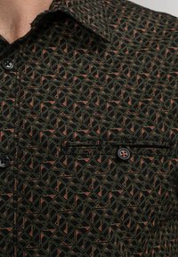 Korte mouw shirt van zwarte stof met een geometrisch patroon van groene en rode driehoeken. Bevat een button-down kraag en een borstzak.