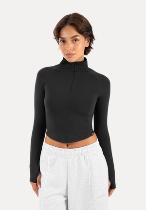 Frau trägt ein schwarzes Langarm-Cropped-Top mit Reißverschluss vorne und hellgraue, hoch taillierte Jogginghose, steht vor weißem Hintergrund.