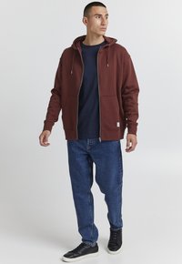 Bordeaux glidelås hettegenser med hette, kombinert med en marineblå skjorte, blå jeans og svarte sneakers. Har lommer foran og snor til justering.