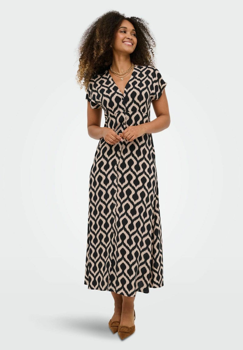 Femme aux cheveux bouclés portant une robe midi à motifs noirs et beiges et des chaussures pointues marron, souriante et les mains jointes.