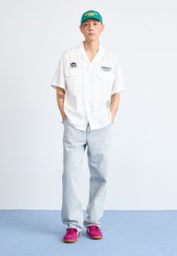Chemise blanche à manches courtes avec deux poches, pantalon en denim bleu clair et baskets roses, complété par une casquette verte avec des logos de marque.