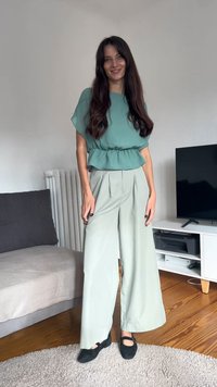 Gaiši zaļa pepluma blūze ar īsām piedurknēm, saskaņota ar platām gaiši zaļām biksēm. Melni apavi un vienkāršs, neitrāls fons.