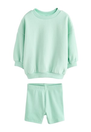 Lille barn med krøllet hår iført lyseblå sweatshirt og shorts står på en græsmark med vilde blomster under en klar himmel.