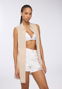 Abito camicia senza maniche beige, realizzato in tessuto leggero, con chiusura a bottoni sul davanti, abbinato a pantaloni corti in denim bianchi strappati e un reggiseno in pizzo bianco.