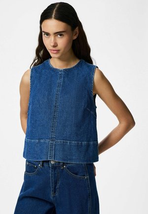 Młoda kobieta o długich ciemnych włosach, ubrana w bezrękawnik z denimu i pasujące jeansy, stoi z rękami za plecami.