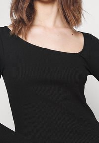 Haut noir à manches longues côtelé avec un col asymétrique. Il présente un tissu texturé et une coupe cintrée, mettant l'accent sur le confort et le style.