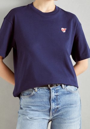 Granatowy bawełniany t-shirt z krótkimi rękawami, okrągłym dekoltem i małym czerwonym haftowanym logo na lewej piersi. Noszony z jasnoniebieskimi dżinsami.