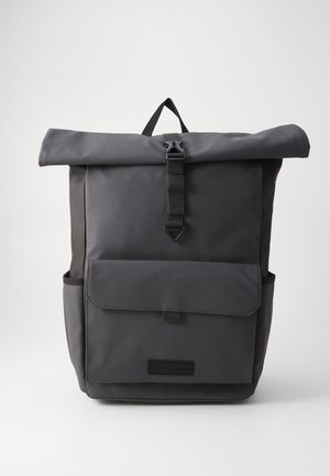 Sac à dos noir à enroulement avec poche avant à rabat, poches latérales et poignée supérieure sur un fond clair uni.