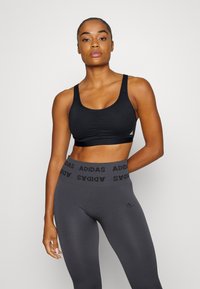 Crni sportski grudnjak s izrezom u obliku supe i podstavljenim čašama, u kombinaciji s visokim sivim tajicama koje na struku imaju ponovljeni crni logo "ADIDAS".