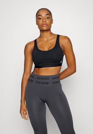 Svart sport BH med rund hals og foret kopper, parret med høy midje grå leggings som har et gjentatt svart "ADIDAS"-logo på midjebåndet.