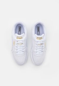 Ett par vita Puma-sneakers med guldaccenter, snörade, sett ovanifrån på en vit bakgrund.