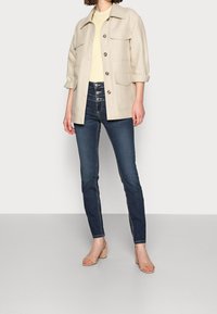 Béžová pracovní bunda s dlouhými rukávy a čtyřmi kapsami, zkombinovaná s tmavě modrými úzkými džíny a skořicovými sandály na blokovém podpatku.