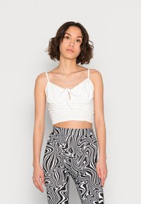 Top curto branco com textura franzida e alças spaghetti ajustáveis, combinado com calças com padrão mármore preto e branco.