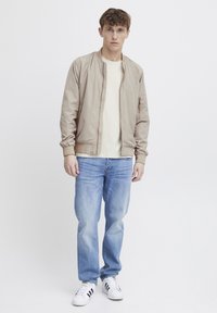 Lys beige bomberjakke, mykt stoff, ribbede mansjetter, glidelåslomme, brukt over en kremfarget T-skjorte, sammen med lyseblå jeans og hvite sneakers.