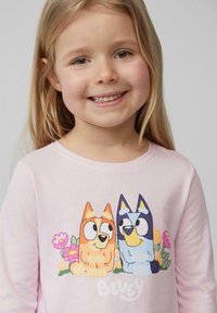 Lichtroze shirt met lange mouwen met cartoonhonden. Het ontwerp bevat oranje en blauwe figuren omringd door kleurrijke bloemen.