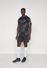 T-shirt de sport noir avec un motif subtil, logo de Tottenham et marque Nike ; associé à des shorts foncés, des chaussettes blanches et des crampons noirs.
