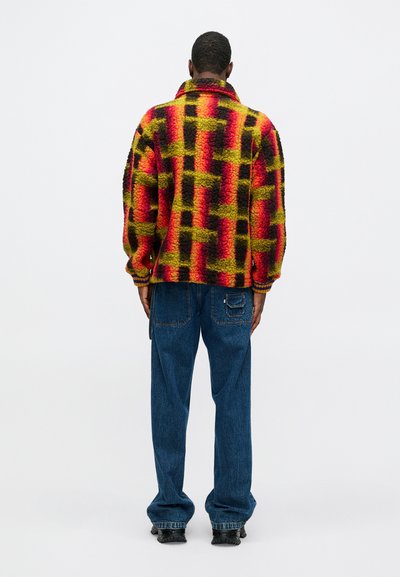 Vilkšninė striukė ryškiai raudonos, geltonos ir juodos patikros rašto; turi apykaklę ir raibotus rankogalius, dėvima ant mėlynų džinsų.