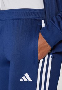 Pantaloni tuta blu navy realizzati in tessuto morbido, con strisce laterali bianche e logo Adidas; include una tasca laterale con cerniera.