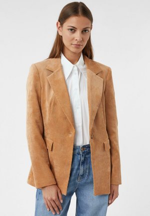 Marynarka ze sztruksu w jasnobrązowym kolorze, z klapami z wycięciem, dwoma przednimi kieszeniami i pojedynczym zapięciem na guzik, zestawiona z białą koszulą i niebieskimi dżinsami.