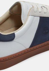 Zapatilla blanca con paneles de ante azul marino, forro interior beige, cordones blancos y suela de goma marrón, con el logo del árbol de Timberland.