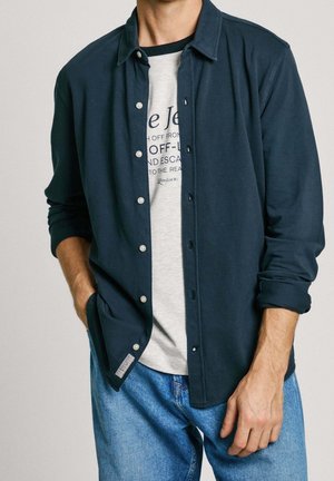 Mężczyzna w rozpiętej ciemnoniebieskiej koszuli z długim rękawem nałożonej na jasnoszarą koszulkę z nadrukiem i niebieskie dżinsy, z jedną ręką w kieszeni.