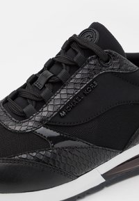 Gros plan d'une basket noire Michael Kors avec motif peau de serpent texturé, tissu en mesh et logo de marque sur le côté et la languette.