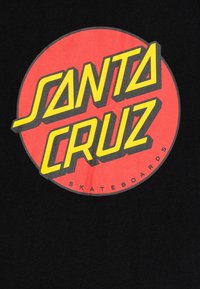 Rund logo med "SANTA CRUZ" i fet gul tekst på rød bakgrunn, omkranset av en svart sirkulær kant, på svart stoff.