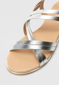Sandales métalliques argentées avec des lanières croisées et une semelle en cuir beige. La semelle est lisse et la finition a une texture subtile.