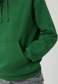 Un hoodie vert en tissu doux, avec une poche avant et des poignets côtelés. Le design comprend une capuche avec cordon de serrage et une coupe décontractée.