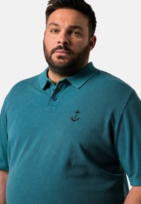 Tyrkysové polo tričko vyrobené z měkké látky, s klasickým límečkem a hnědou vyšitou kotvou na levé části hrudi. Krátké rukávy, volný střih.