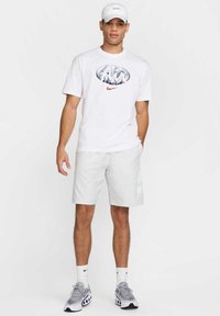 T-shirt blanche avec un logo Nike argenté, accompagnée de shorts gris clair dotés d'un logo Nike décoloré, portée avec des chaussures de sport grises et une casquette blanche.