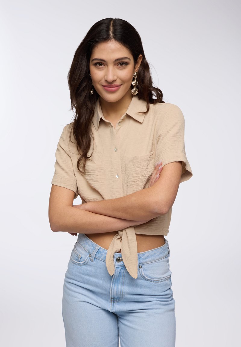 Camicia beige a maniche corte con tessuto testurizzato e chiusura a bottoni, legata in vita. Indossata con jeans a vita alta di colore azzurro chiaro.
