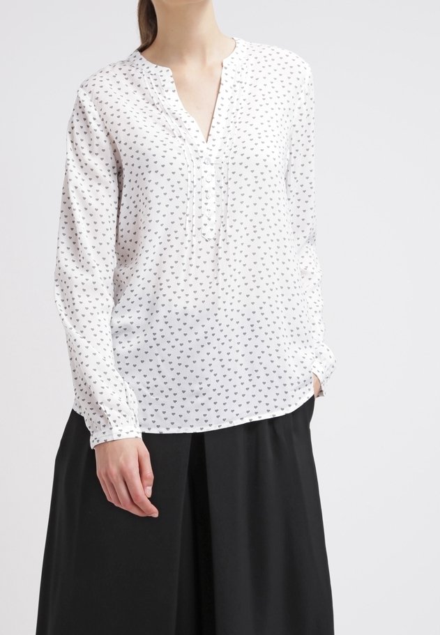 Valkoinen pitkähihaisuus paita, jossa on V-pääntie ja pieni musta sydäntyyli, nappi ranteissa ja löysä istuvuus. Yhdistetty mustaan hameeseen.