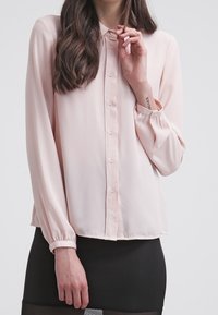 Blusa rosa chiara realizzata in un tessuto morbido, con chiusura a bottoni sul davanti, maniche lunghe con polsini elasticizzati e una vestibilità morbida, indossata con una gonna nera.