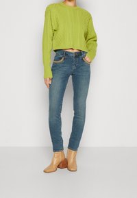 Suéter corto de punto de cable en verde lima, jeans ajustados azul de tiro medio con bolsillos decorativos y botas tobilleras de ante beige con tacones cuadrados.