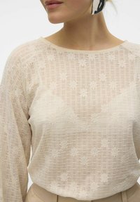 Lys beige bluse lavet af gennemsigtig lace-stof med et gittermønster og blomster motiver. Rund halsudskæring og lange ærmer.
