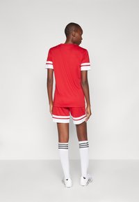 Červený športový dres a šortky s bielymi manžetami a pruhmi. Spárované s bielymi ponožkami po kolená so čiernymi pruhmi a futbalovými kopačkami.