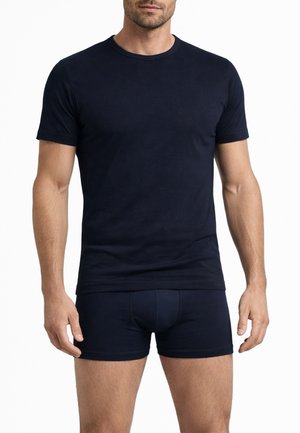 Uomo che indossa una maglietta nera aderente a maniche corte e boxer neri coordinati, in piedi su uno sfondo bianco.