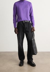 Man som bär en lila zip-up-tröja, svarta jeans, svart bälte och svarta skor, hållande en svart läderhandväska vid sin sida.